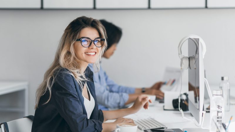Attractive laughing freelancer girl posing with cup of coffee at her workplace. Chinese student in blue shirt works with document in campus with blonde friend in glasses.. Cursos Especializados