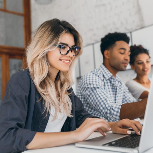 Beautiful curly female freelancer with cute manicure using laptop and smiling. Indoor portrait of blonde secretary sitting beside african coworker in blue shirt.. Programa de Beneficios