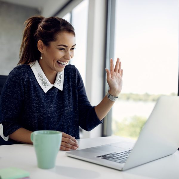 Cheerful businesswoman waving during video call in the office. Happy businesswoman using laptop and having fun while greeting someone during a video call in the office.