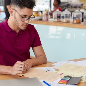 Image of focused male student prepares report in finances, looks attentively at papers, eats delicious croissants, poses over cafe interior with free space for your promotion. Freelance work Vacantes