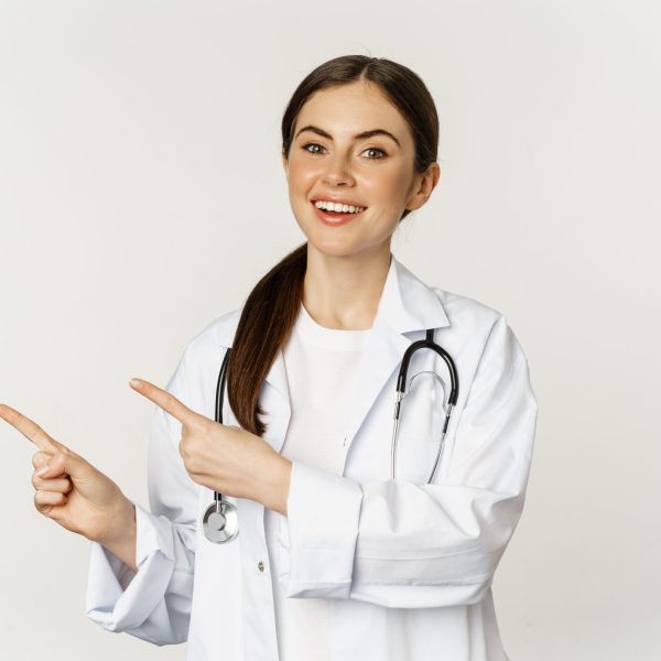Portrait of smiling young woman doctor, healthcare medical worker, pointing fingers left, showing clinic promo, logo or banner, standing over white background Diplomado en Neurociencias